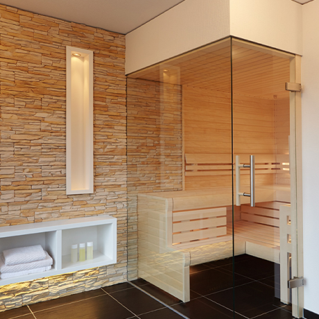Heller Wellnessraum mit Glas-Sauna aus Holz, Natursteinwand, weißen Regalen und indirekter Beleuchtung